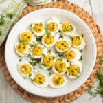 Deviled eggs with curry and dill in a white serving plate on a bamboo placemat on a white table with fresh dill scattered and a green and white plaid towel.