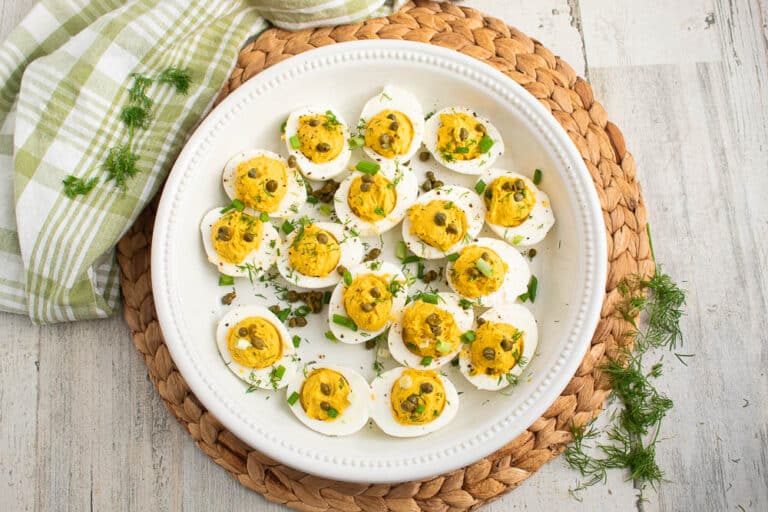 Deviled eggs with curry and dill in a white serving plate on a bamboo placemat on a white table with fresh dill scattered and a green and white plaid towel.