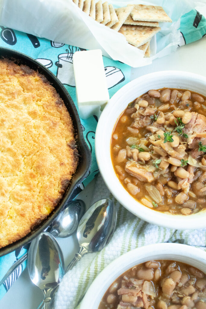 White bowlful of pinto beans and ham along side Easy Cast Iron Skillet cornbread in a black cast iron skillet. Butter and crackers on the side.