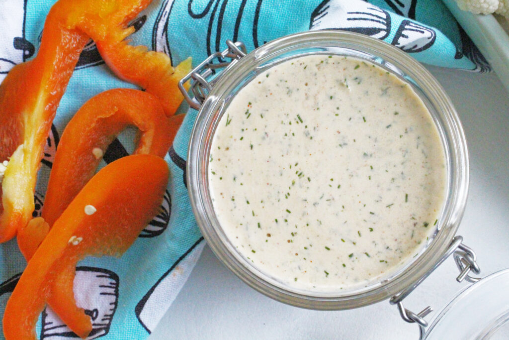 Close up overhead shot of jalapeno chipotle ranch dip in a glass jar with orange bell peppers on a teal towel.