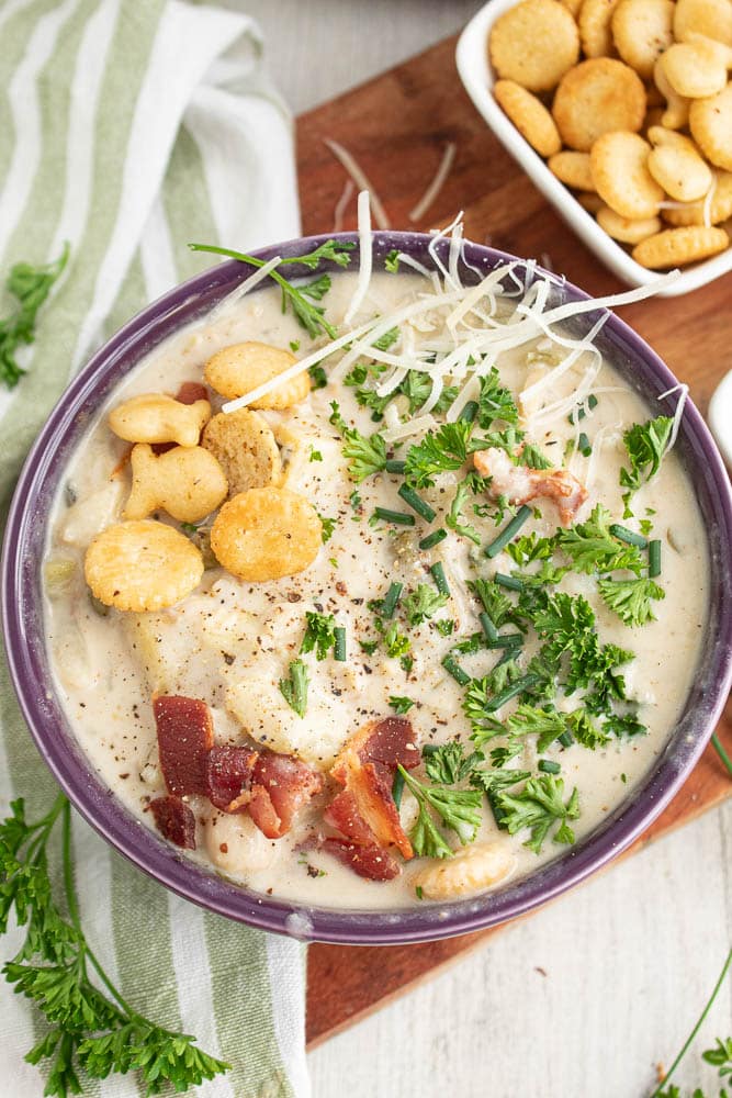 bowl of light clam chowder with oyster crackers bacon chives parsley in a purple bowl