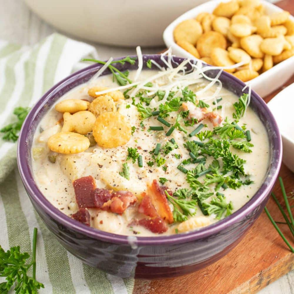 light new england clam chowder with oyster crackers, bacon, chives and parsley in a purple bowl