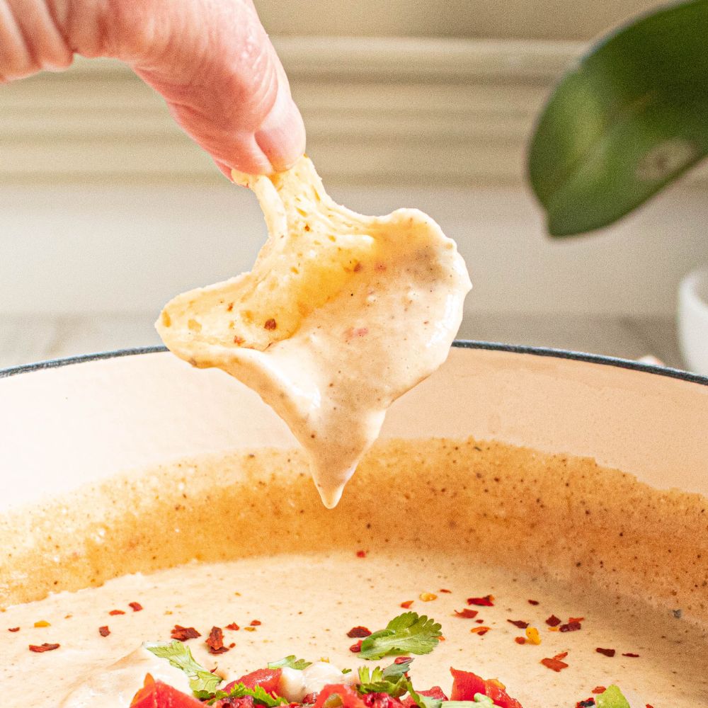 Light and creamy white cauliflower queso dip on a white tortilla chips dripping into the bowl.
