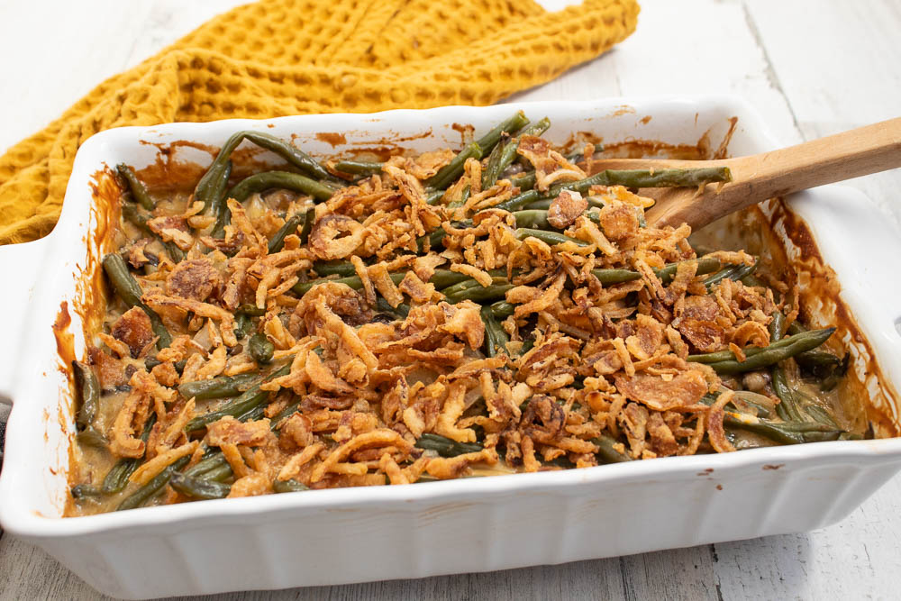 Green Bean Casserole with water chestnuts in a rectanble white casserole dish with a wood serving spoon and mustard yellow towel.