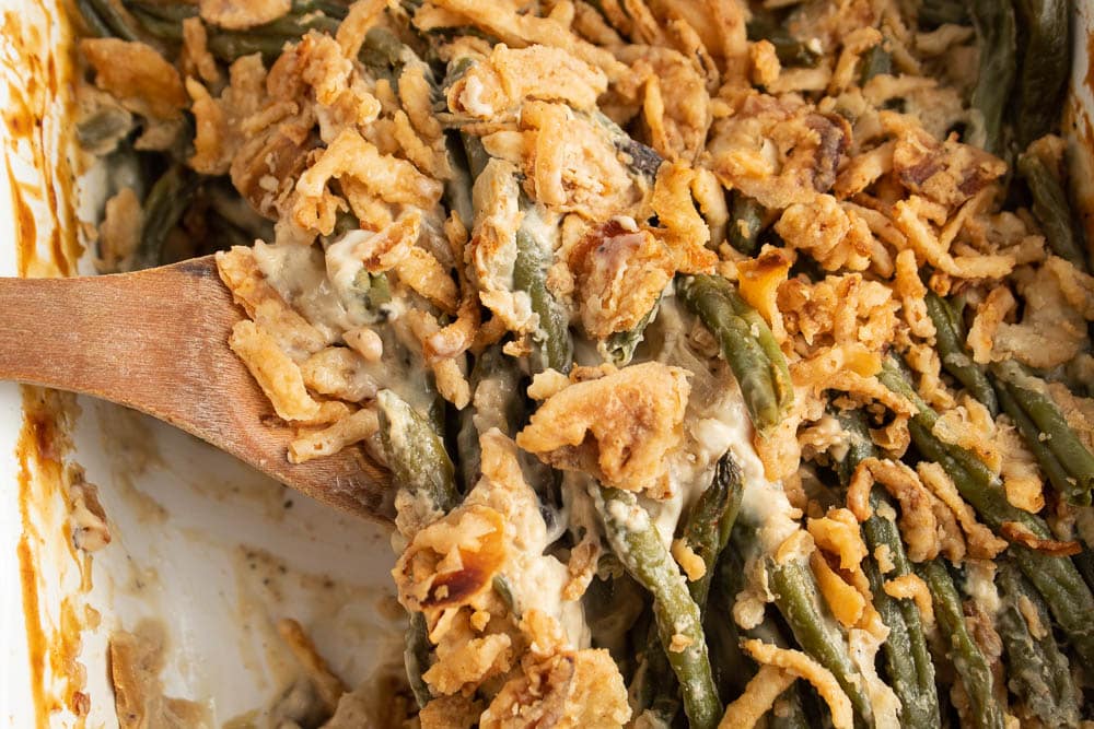 Cooked green bean casserole in a white rectangle casserole dish with baked canned french fried onions on top with a wood serving spoon.