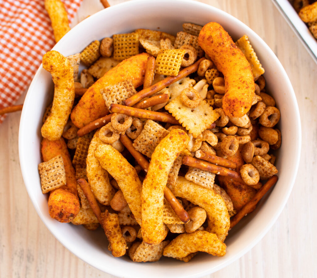 spicy chex mix with cheese puffs in a white bowl on a white table with an orange towel.