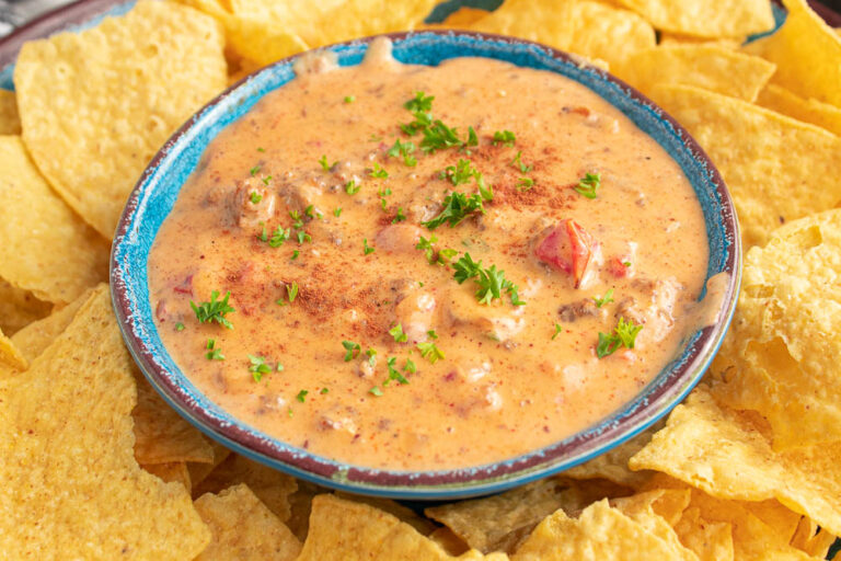 Bowl full of spicy sausage Rotel Queso Cheese Dip in a blue bowl surrounded by yellow tortilla chips.