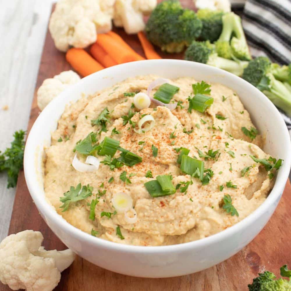 Basic Homemade Hummus Dip in a white bowl with assorted vegetables on a brown serving board.