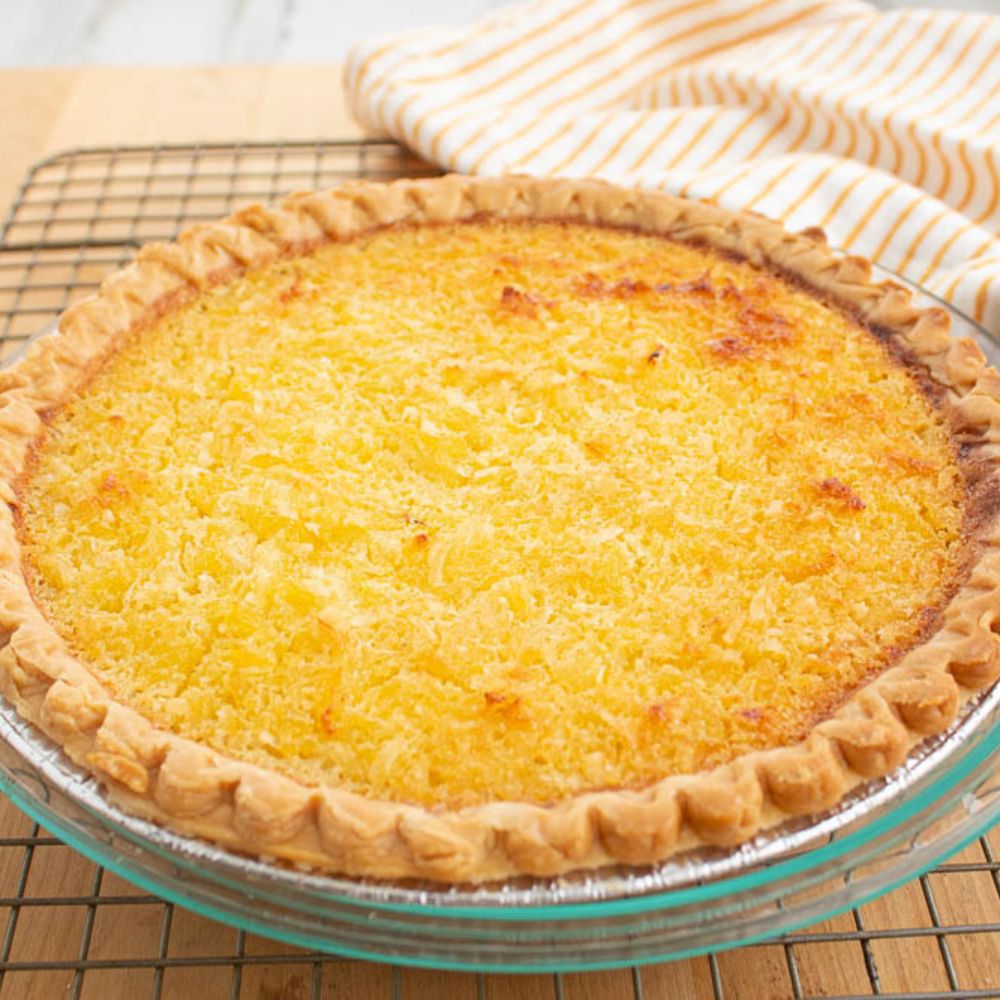 Whole creamy Pineapple Coconut Pie in a clear pie dish on a baking rack with a orange and white towel.
